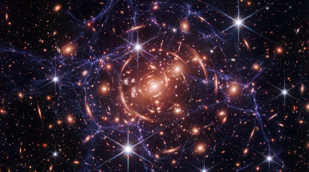 A deep space field filled with colorful galaxies, warped light arcs, and faint web-like filaments connecting star clusters.