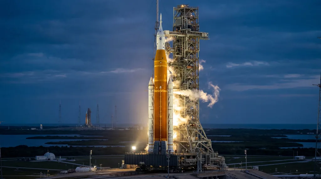 Massive SLS rocket on a launch pad venting white steam plumes during sunrise at Kennedy Space Center.