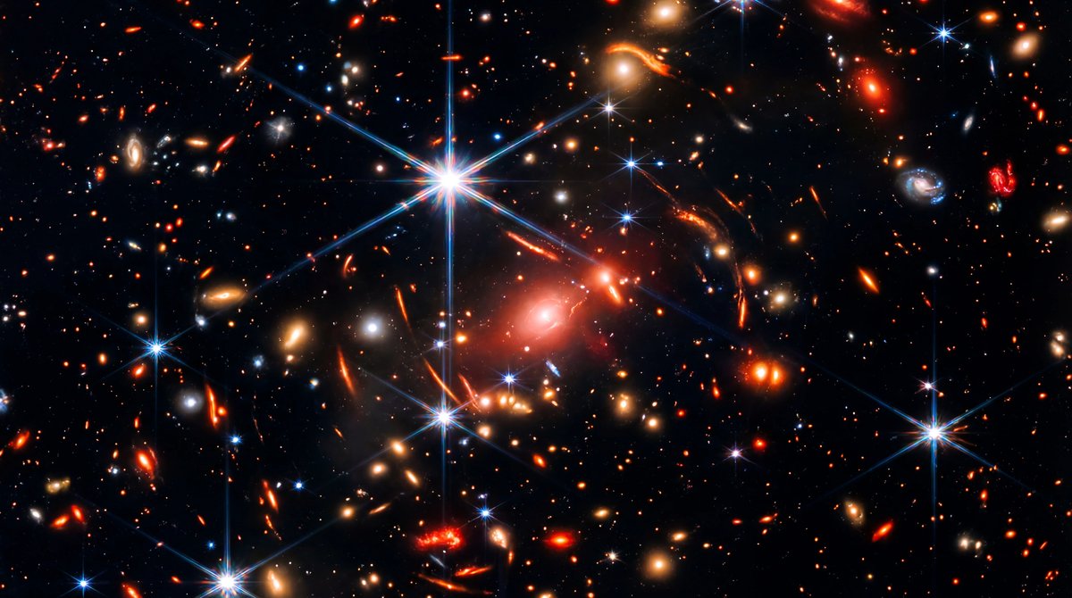A deep space field filled with colorful spiraling galaxies and bright stars with six-pointed spikes, highlighting a small red dot.
