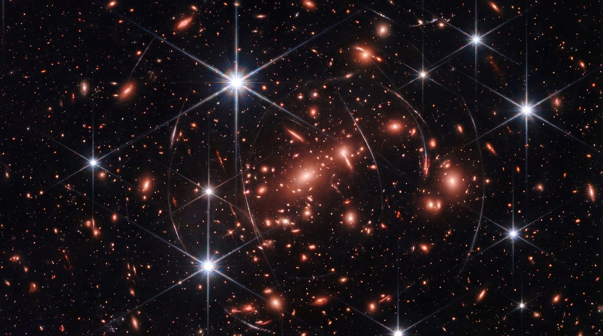 Deep space image filled with thousands of galaxies, highlighting faint red ancient galaxies and bright stars with spikes.