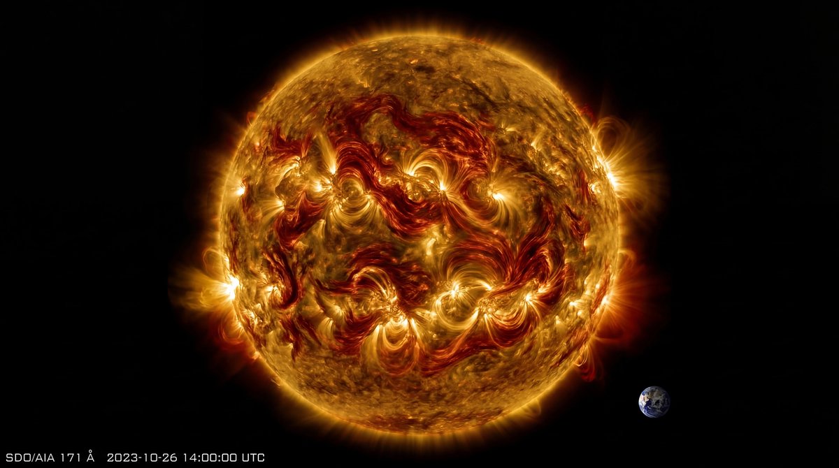 Burning orange Sun with swirling magnetic lines and plasma loops, set against black space with a tiny Earth for scale.